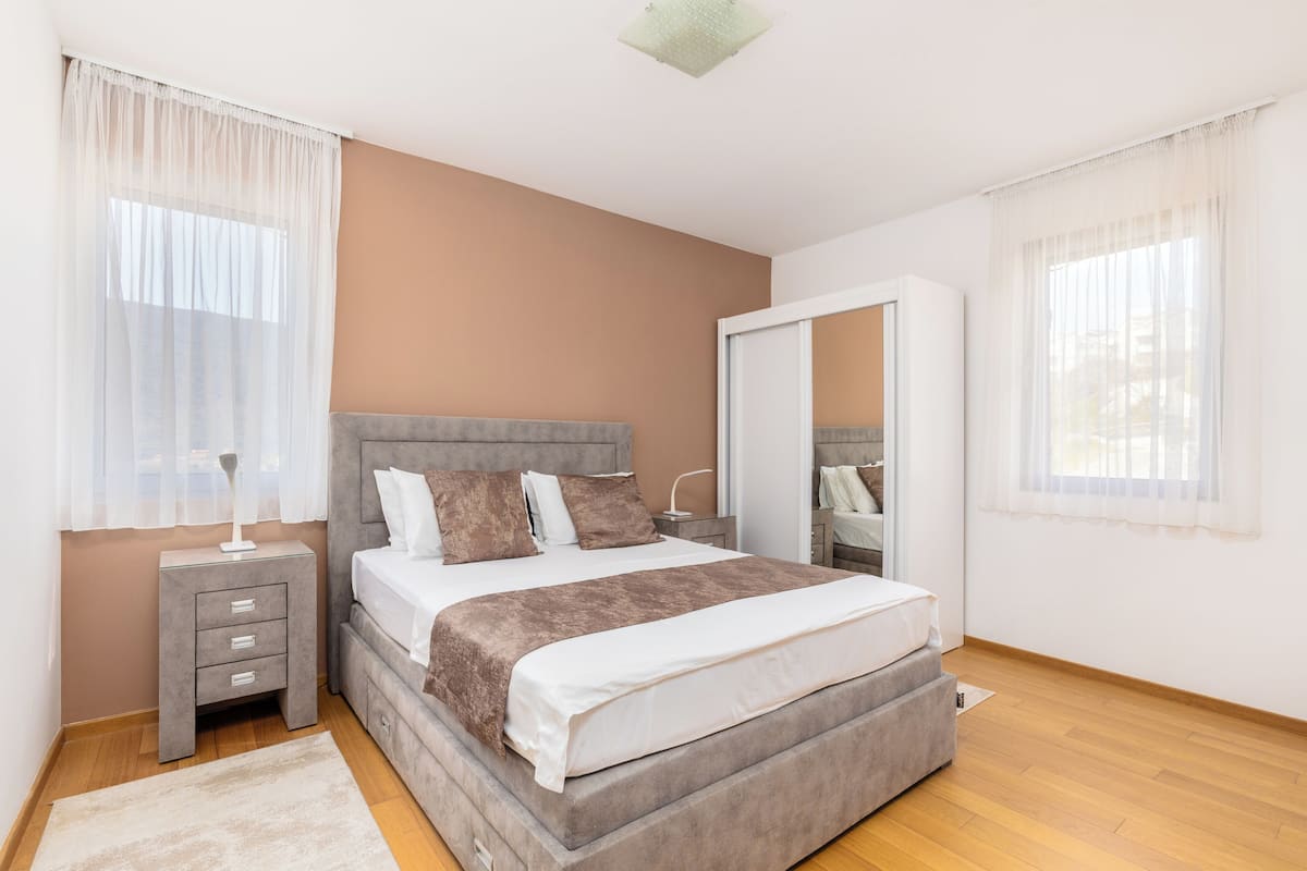 Ein Schlafzimmer mit Bett, Nachttisch, Kommode, Spiegel und einem Fenster mit leichtem Vorhang.
