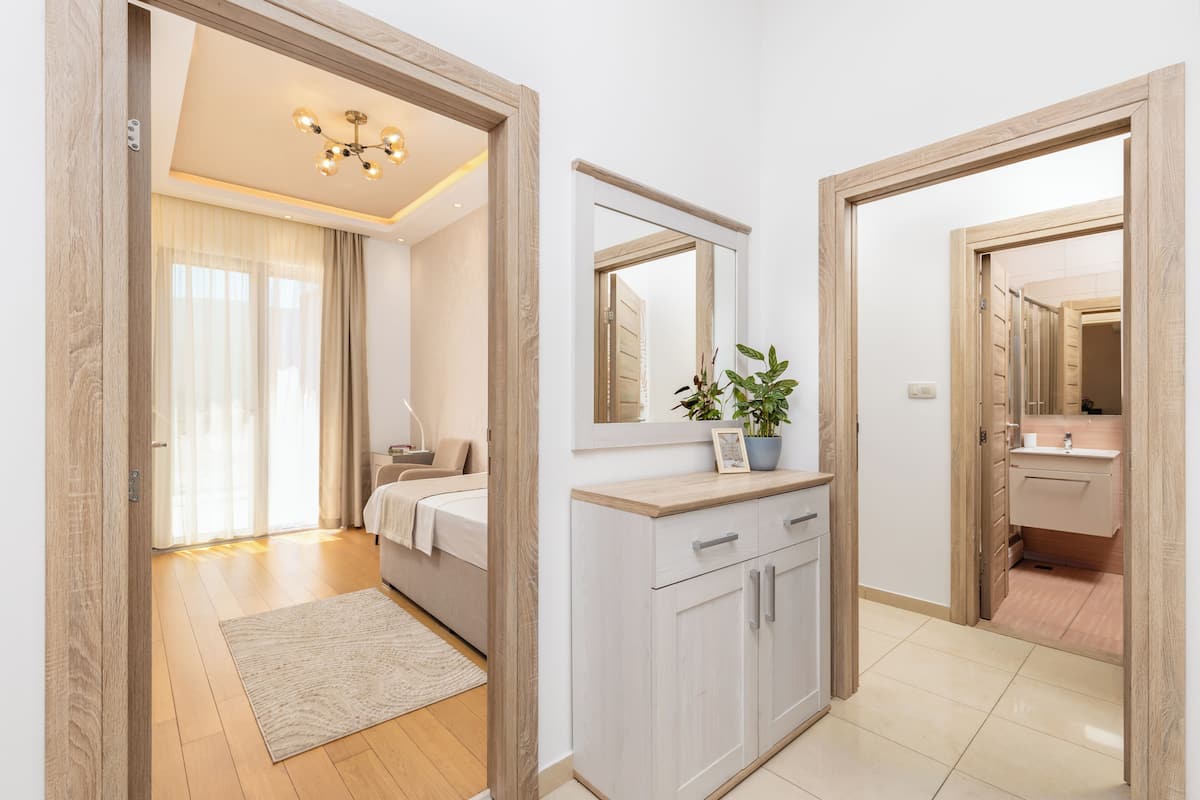 Ein modernes Schlafzimmer mit Bett, Nachttisch und großem Spiegel. Es gibt ein Badezimmer mit Waschbecken und Spiegel. Der Raum hat Holzboden und einen hellen Teppich.
