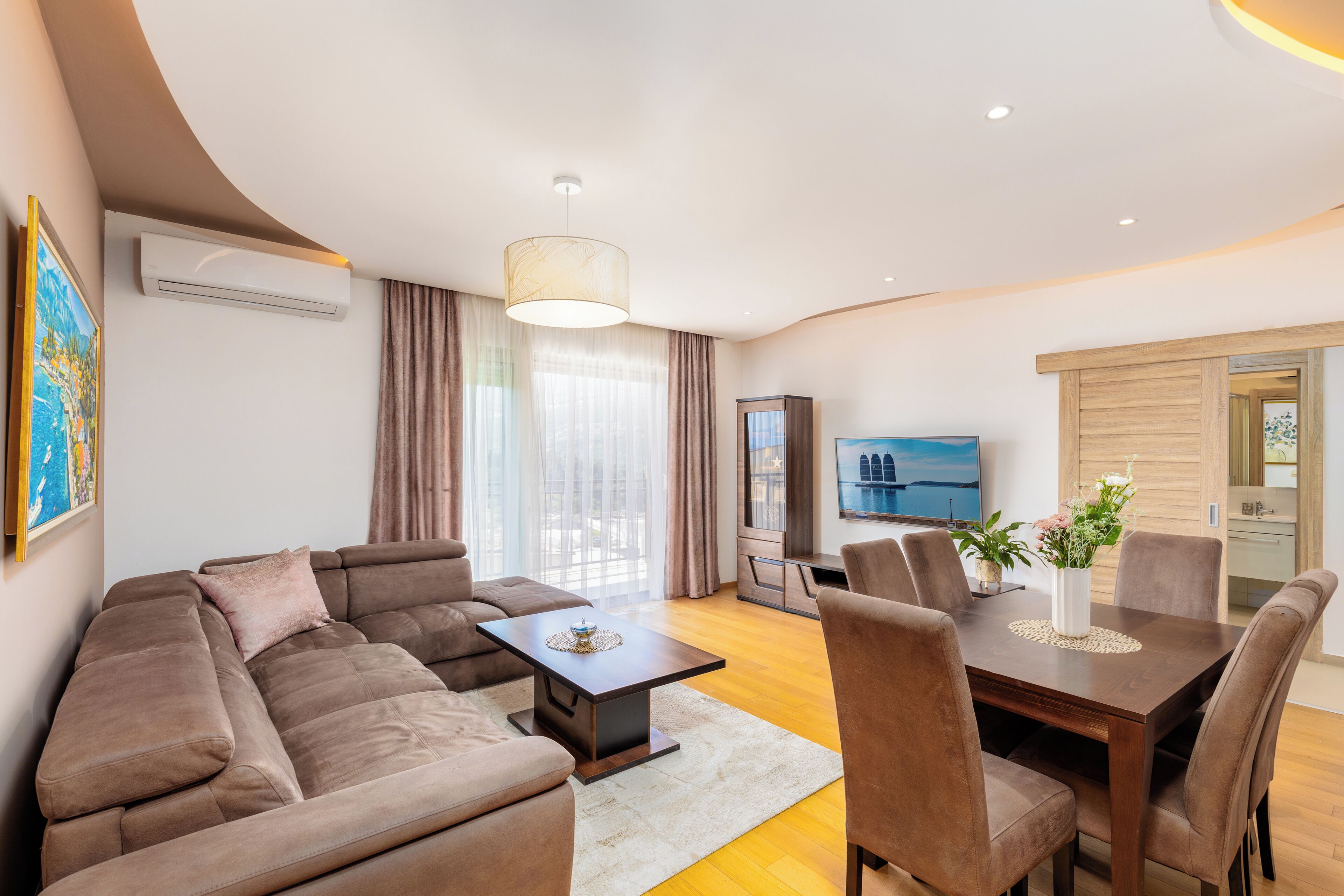 A modern living room with a sofa, a dining table with chairs, a TV, and a large window with curtains.