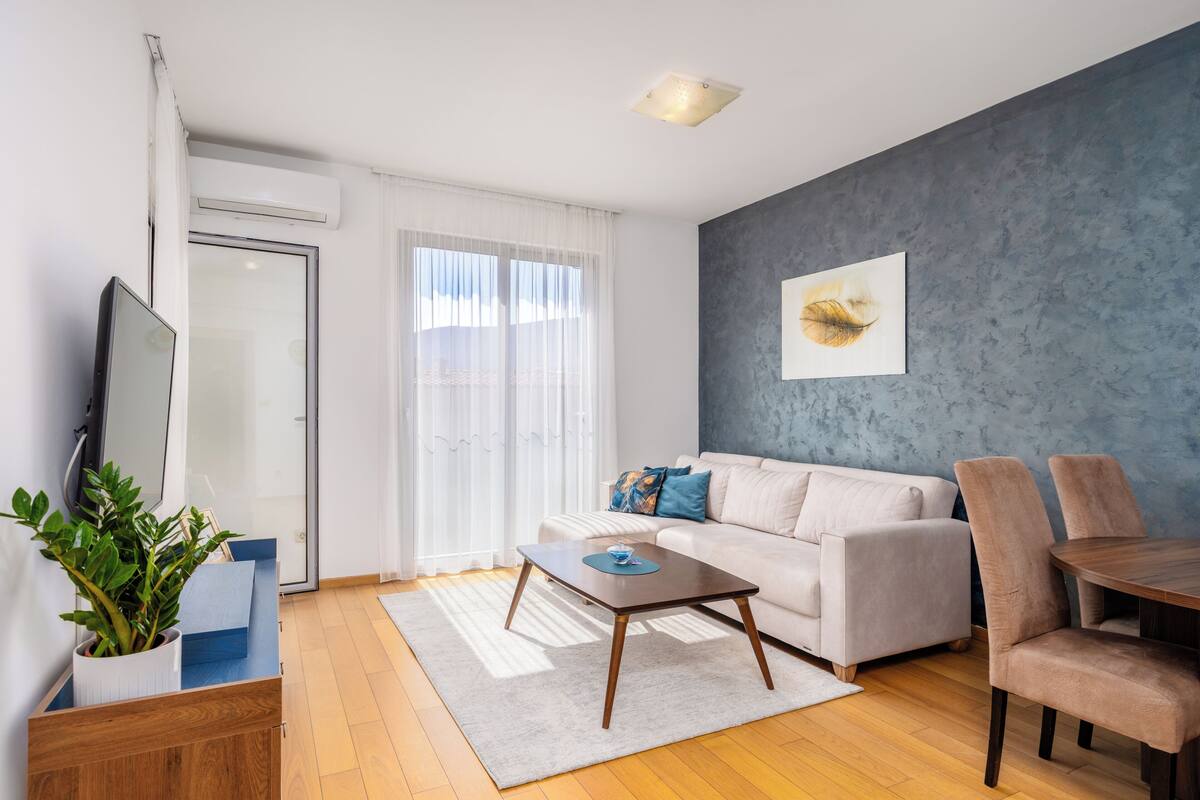 Ein modernes Wohnzimmer mit einer Couch, einem Couchtisch und Sesseln, einem Flachbildfernseher und einem großen Fenster mit Vorhängen.