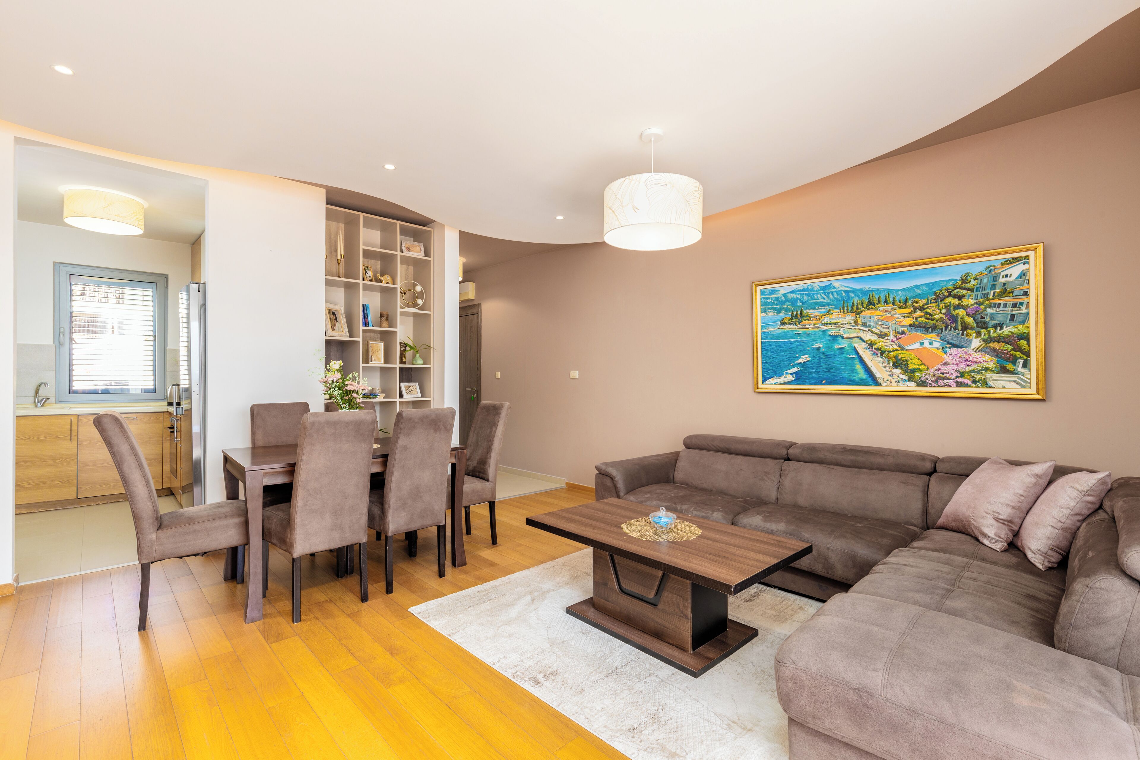 A modern living room with a dining area, a large sofa, and a painting of a coastal town.