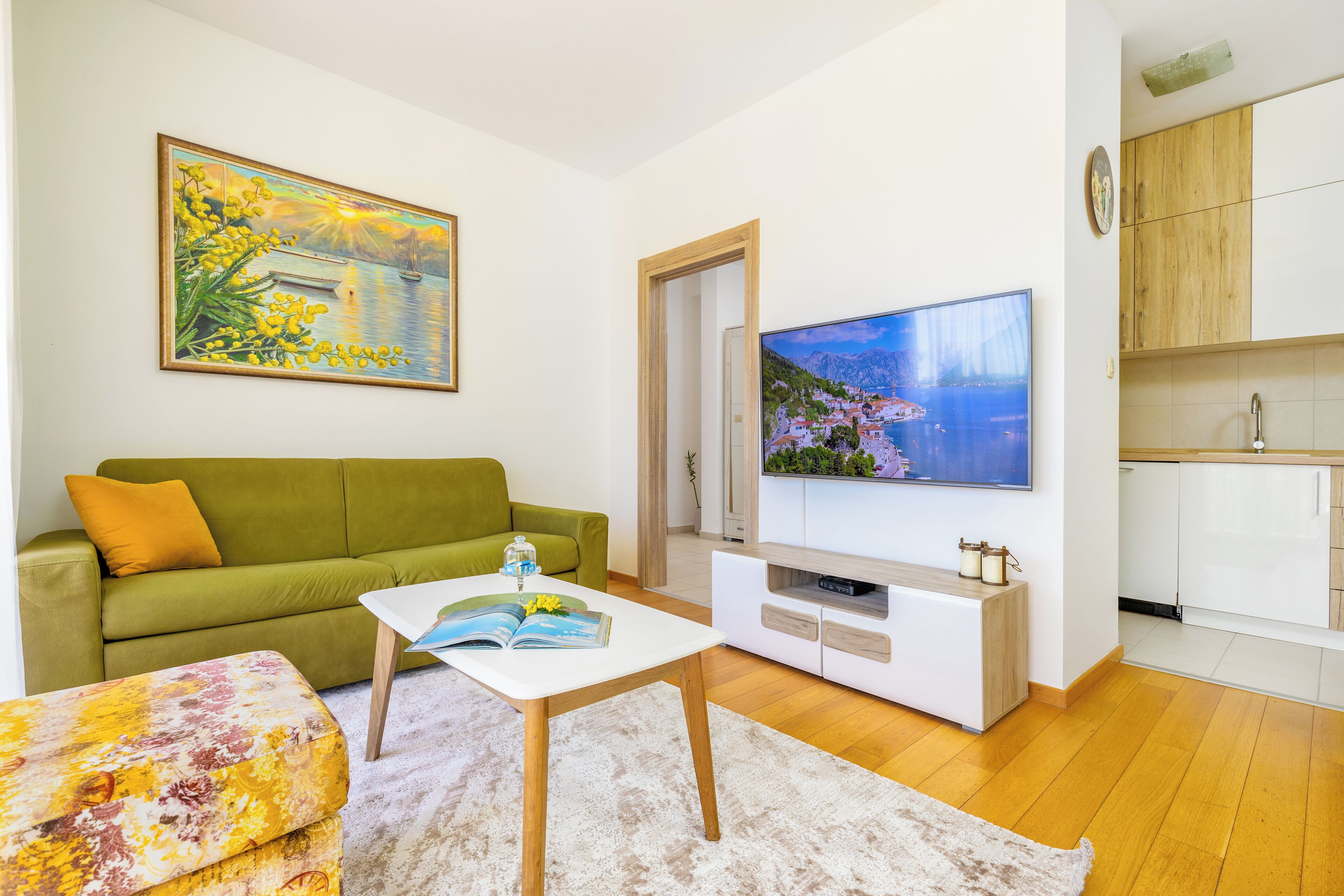 Una sala de estar con un sofá verde, una mesa de centro blanca, un cuadro de una escena costera y un televisor que muestra un paisaje pintoresco.
