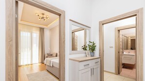 Een moderne slaapkamer met een bed, nachtkastje en een grote spiegel. Er is een badkamer met een wastafel en spiegel. De kamer heeft houten vloeren en een lichtkleurig vloerkleed.