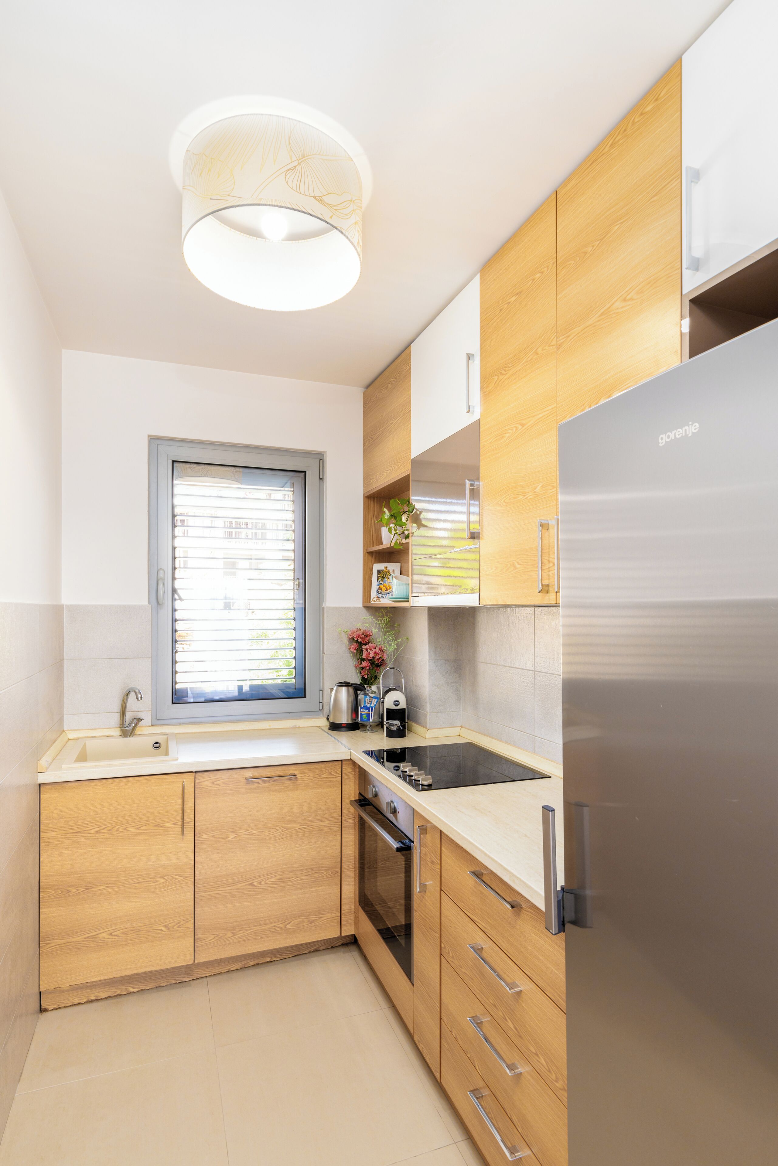 A modern kitchen with wooden cabinets, a stainless steel refrigerator, a built-in oven, and a window with blinds.
