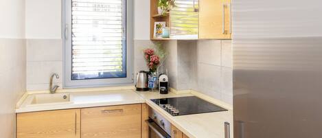 A modern kitchen with wooden cabinets, a stainless steel refrigerator, a built-in oven, and a window with blinds.