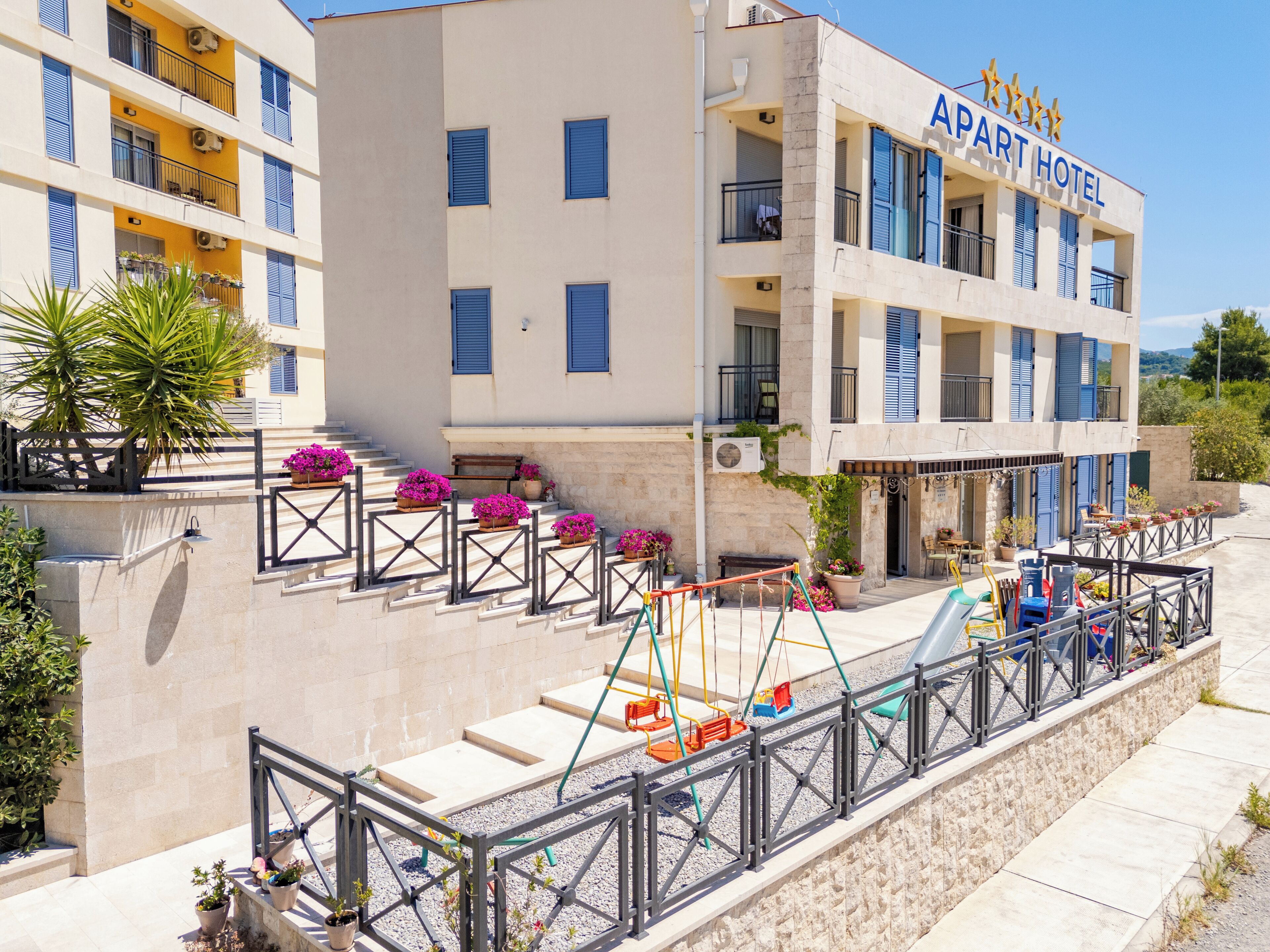 A multi-story building with a sign reading "APART HOTEL". There are balconies with blue shutters, potted plants, and a playground with swings and a slide.