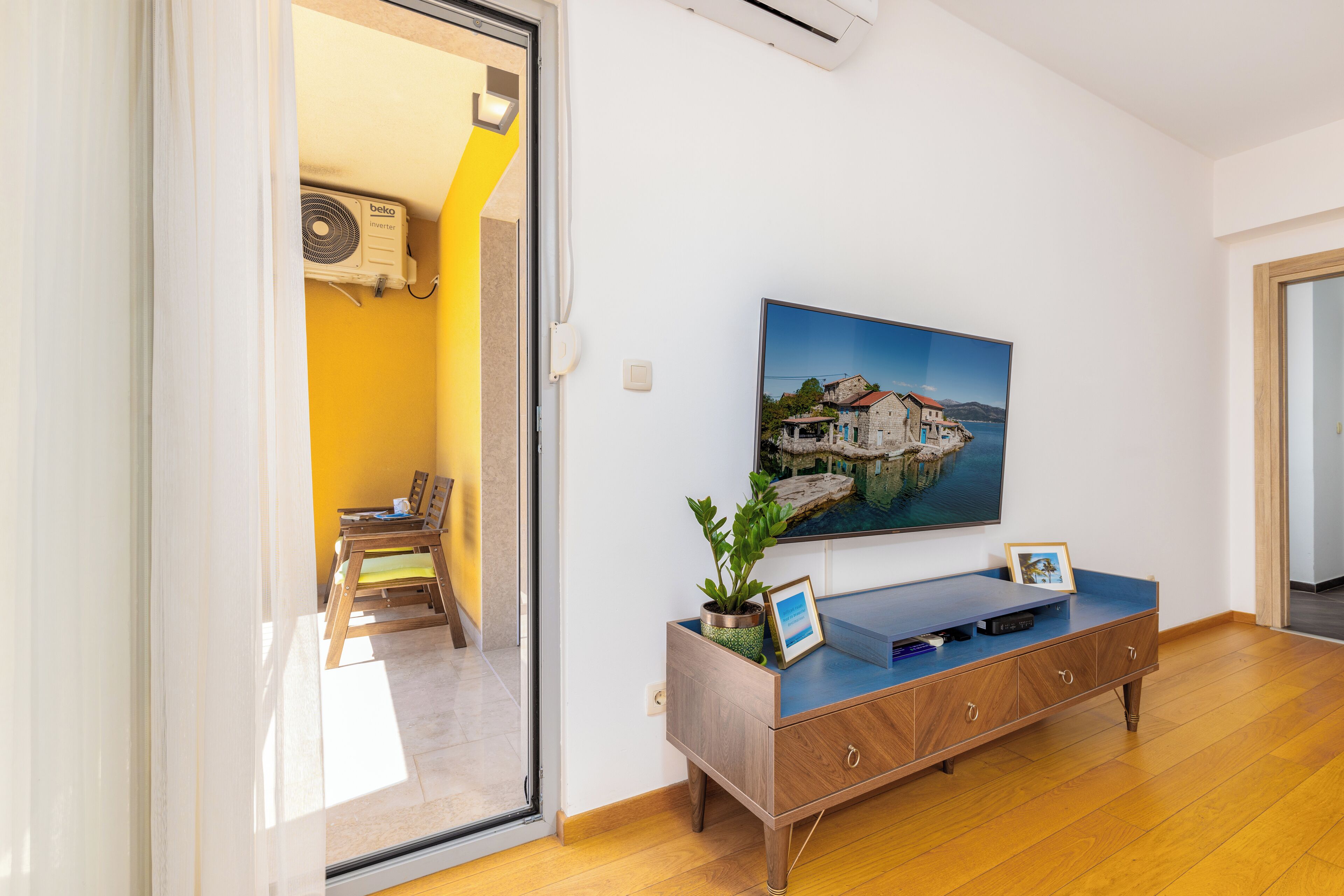 Una habitación con piso de madera, un sofá azul, un televisor en la pared, una planta en maceta y un cuadro.