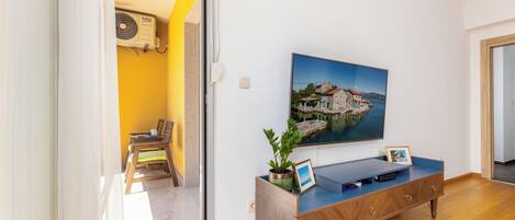 Una habitación con piso de madera, un sofá azul, un televisor en la pared, una planta en maceta y un cuadro.