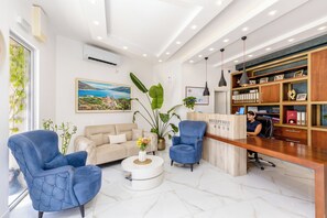 Un espace d’accueil comprenant un comptoir de réception, deux fauteuils bleus, un canapé beige, une table basse ronde ornée de fleurs, une grande plante en pot et une bibliothèque avec des photos encadrées et des classeurs.