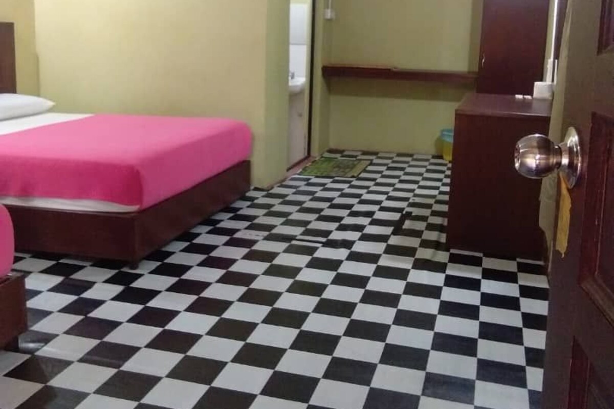 A room with a checkered floor, a bed with a pink bedspread, a wooden dresser, and a door with a round handle.