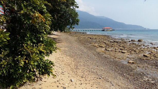 A rocky beach with a pier extending into the water, a red-roofed building, and mountains in the background. - Johan's Place (Tioman Island)