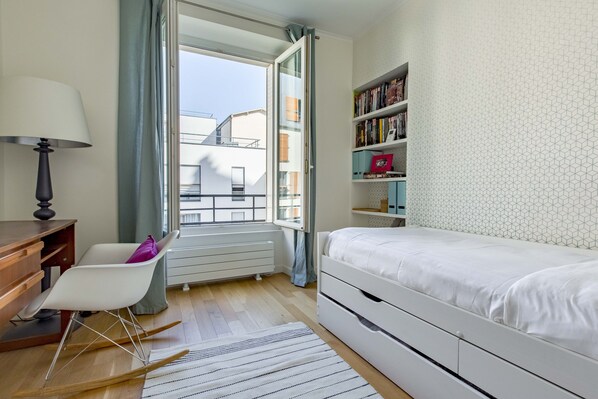 Une chambre à coucher avec un lit blanc, un bureau avec une lampe, un fauteuil à bascule, une bibliothèque et une fenêtre avec des rideaux.