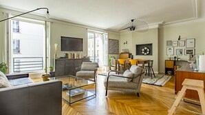 A modern living room with a sofa, armchairs, a glass coffee table, and a fireplace. There is a dining area with a table and chairs, and a kitchen area with a counter and appliances.