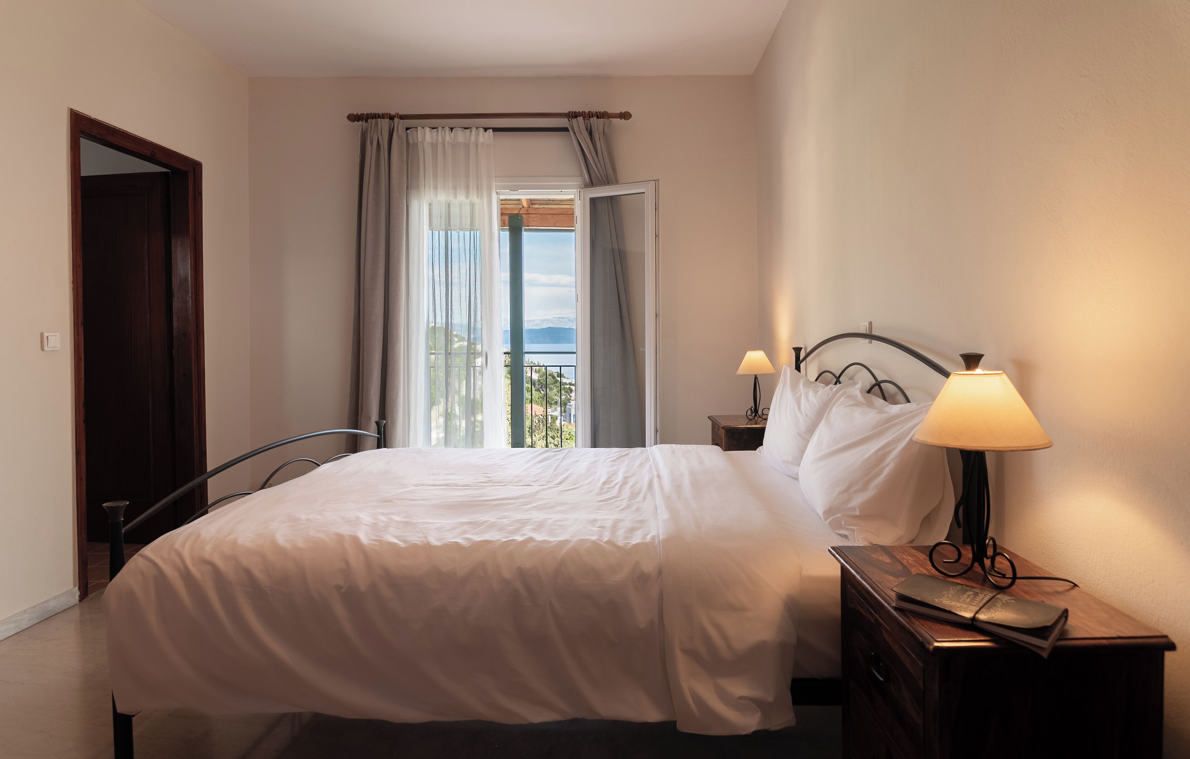 Ein ordentlich bezogenes Bett in einem Zimmer mit Blick ins Freie durch ein Fenster mit Vorhängen. Ein Nachttisch mit Lampe und Buch ist vorhanden.