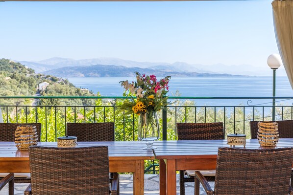 A terrace with wicker furniture, a table with a vase of flowers, and a view of the sea and mountains.