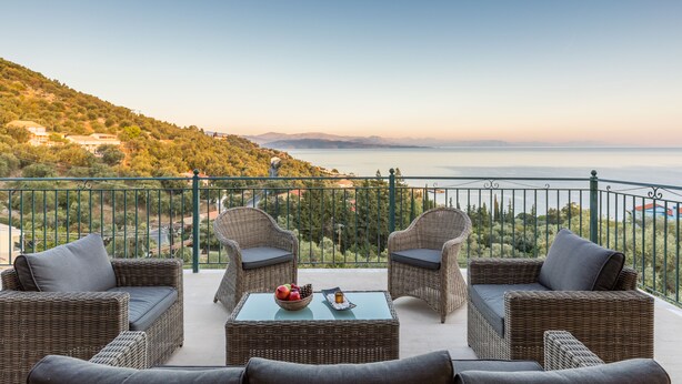 Un balcon agrémenté de meubles en osier, une table avec des fruits et une vue sur la mer et les collines.