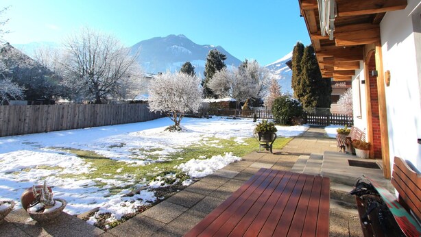 Une terrasse en bois avec un banc, un bac à fleurs en pierre contenant un arbre, et une vue sur une chaîne de montagnes.