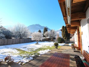 Une terrasse en bois avec un banc, un bac à fleurs en pierre contenant un arbre, et une vue sur une chaîne de montagnes.
