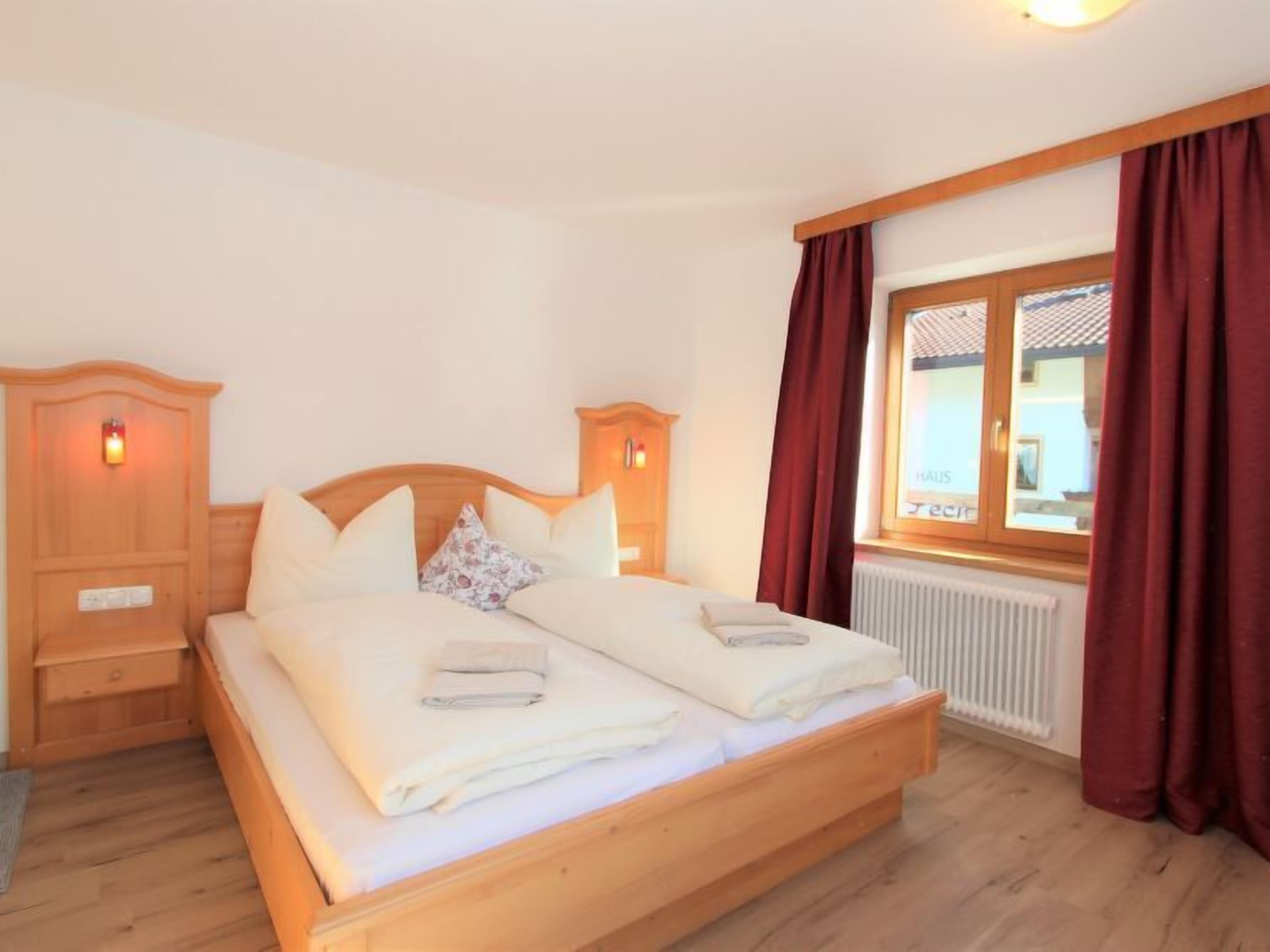 Ein ordentlich bezogenes Doppelbett mit Holz-Kopfteil, ein Nachttisch mit Lampe und ein Fenster mit Vorhängen.