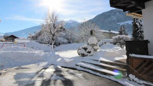 Un paysage enneigé avec une montagne en arrière-plan, un bâtiment en bois doté d’un balcon et un sentier recouvert de neige.