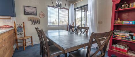 Un comedor con mesa y sillas de madera, una araña y una estantería con juegos de mesa.
