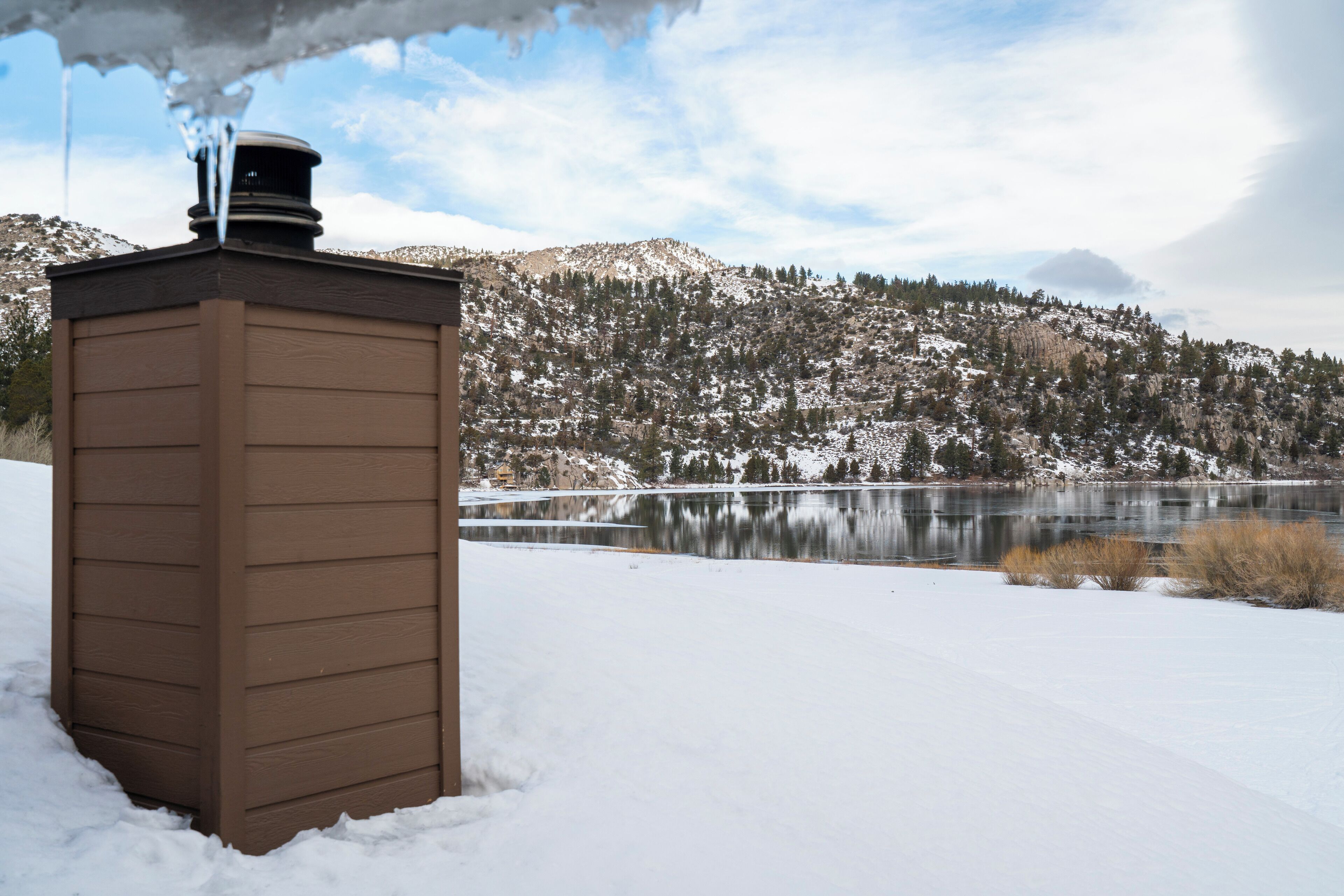 Une cabane en bois avec une cheminée, située près d’un lac, avec des collines enneigées en arrière-plan.