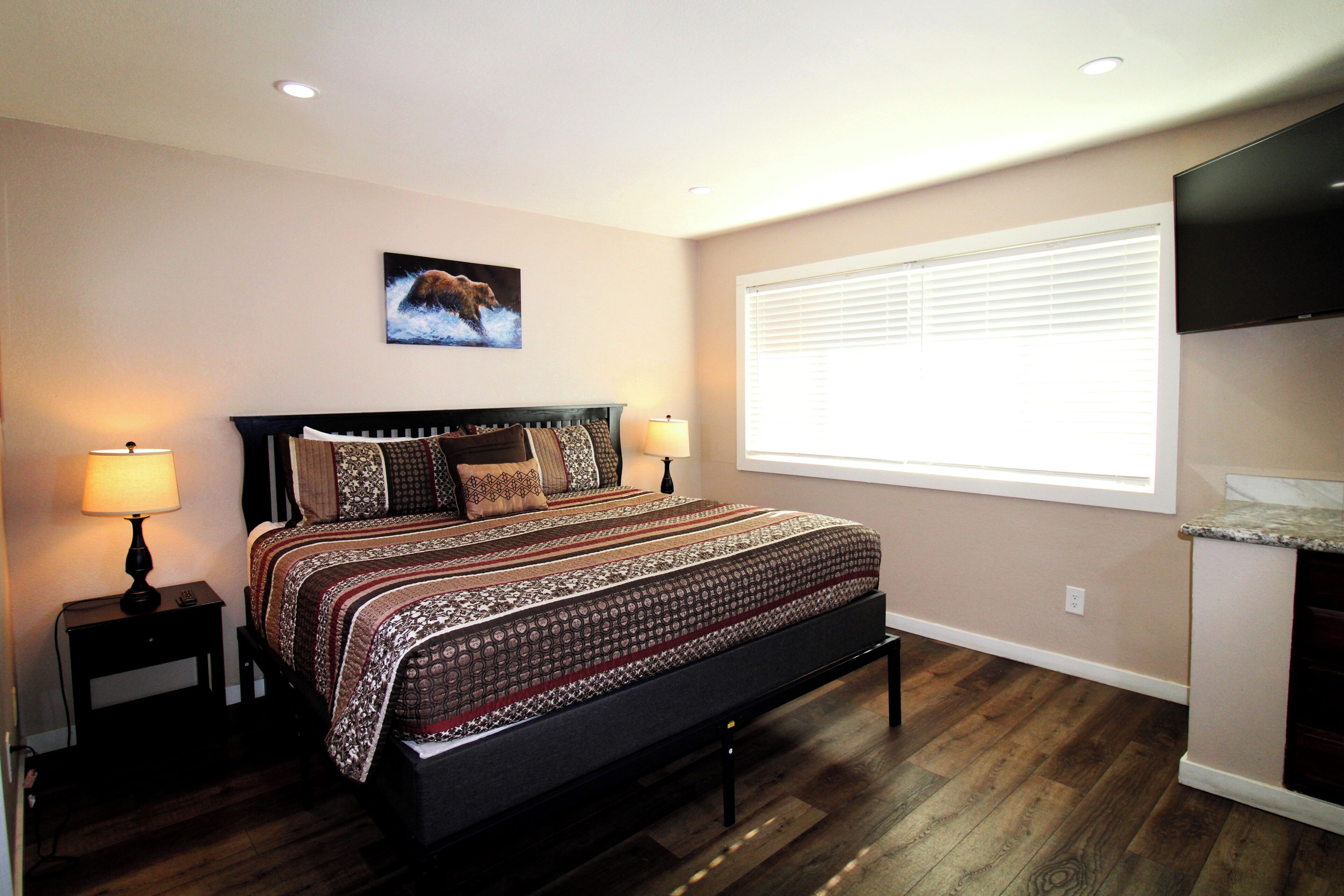 A bedroom with a large bed, two bedside tables with lamps, a wall-mounted TV, and a window with blinds.