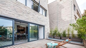 A modern outdoor patio with wooden flooring, a glass door leading to a dining area, and a seating arrangement with two chairs and a small table.