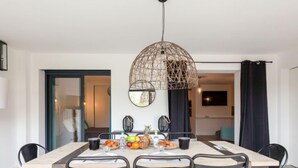 A dining area with a round table, six chairs, a hanging light fixture, and a view into another room.