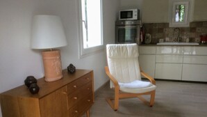 Une cuisine moderne équipée d’un four à micro-ondes, d’un four et d’un évier. On y trouve une commode en bois avec une lampe et des objets décoratifs. Une chaise blanche rembourrée se trouve au premier plan.