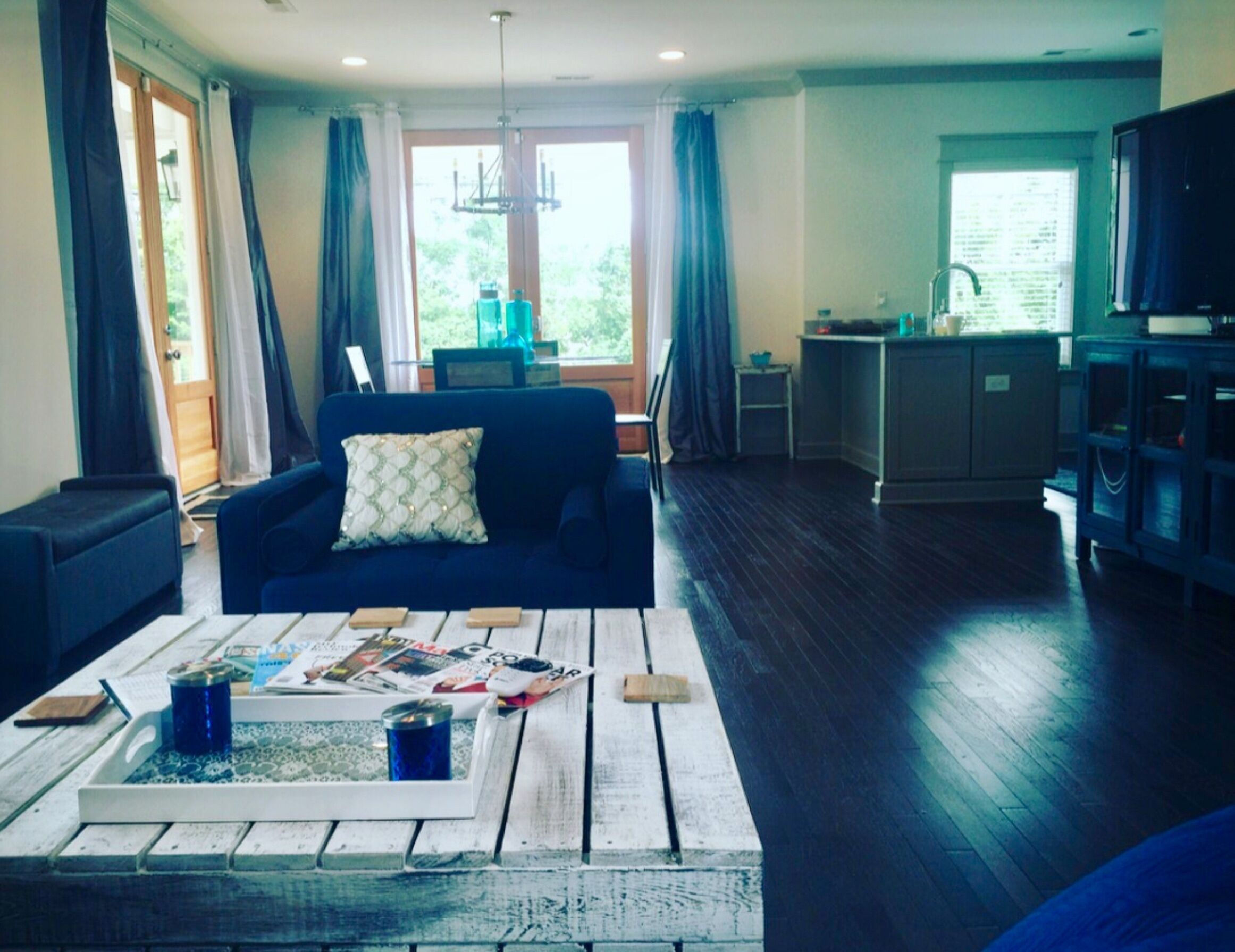 Una sala de estar con un sofá azul, una mesa de centro con revistas y bebidas, una zona de cocina con pileta y armarios, y un ventanal con cortinas.