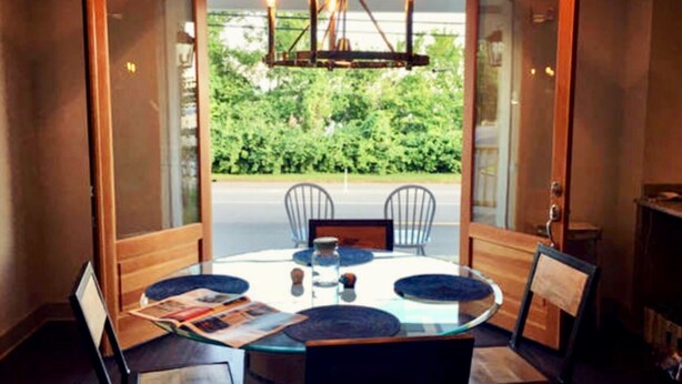 A round glass table with four chairs, a chandelier, and a view of greenery through glass doors.