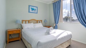 A neatly made bed with white linens, a wooden headboard, and a small bedside table with a lamp. - Castillo Del Mar 10 - Overlooking the Lake (Tuncurry)