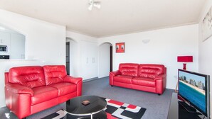 A modern living room with two red leather sofas, a black coffee table, and a flat-screen TV displaying a cityscape. - Albert Court 8 - Acrpss from Wallis Lake! (Forster)