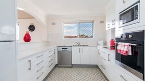 A compact kitchen with white cabinets, a dishwasher, a microwave, and an oven. There's a window with a view of the outside, and a red vase on the counter. - Albert Court 8 - Acrpss from Wallis Lake! (Forster)