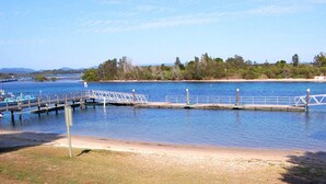 A pier extending into a calm body of water with a sandy beach in the foreground and a forested area in the background. - Albert Court 8 - Acrpss from Wallis Lake! (Forster)