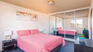 A bedroom with a large bed, a bedside table, a lamp, a mirror, and a view of the outside. - Albert Court 8 - Acrpss from Wallis Lake! (Forster)