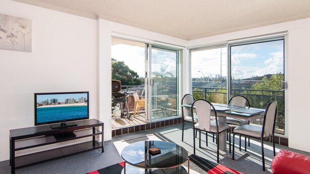 A modern living room with a TV, a glass table, and a balcony with a view of the sea and buildings. - Albert Court 8 - Acrpss from Wallis Lake! (Forster)
