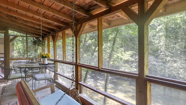 Un área de estar al aire libre techada con una mesa y sillas, rodeada de amplias ventanas que dan a un paisaje boscoso.