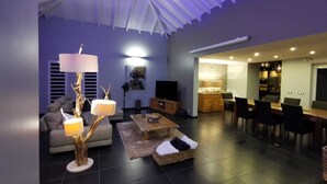 Un salon moderne avec un canapé, une table basse et des lampes décoratives. On y aperçoit une télévision, un coin repas avec des chaises et, au fond, une cuisine.