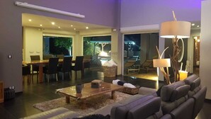 Un salon moderne avec un canapé, une table basse et une lampe. On y trouve également un coin repas avec des chaises et une table. La pièce est dotée de grandes fenêtres et offre une vue sur l’extérieur.