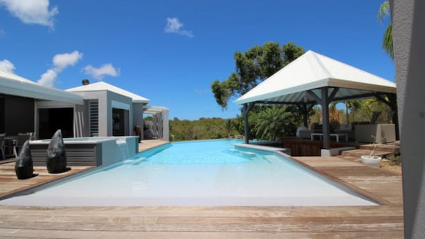 Une piscine avec une terrasse en bois, un espace salon couvert et un bâtiment moderne doté d’un patio.