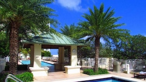 A pool area with a covered seating area, palm trees, and a clear blue sky. - One bedroom in amazing luxury condo. BEACH PASS INCLUDED! (Boynton Beach)