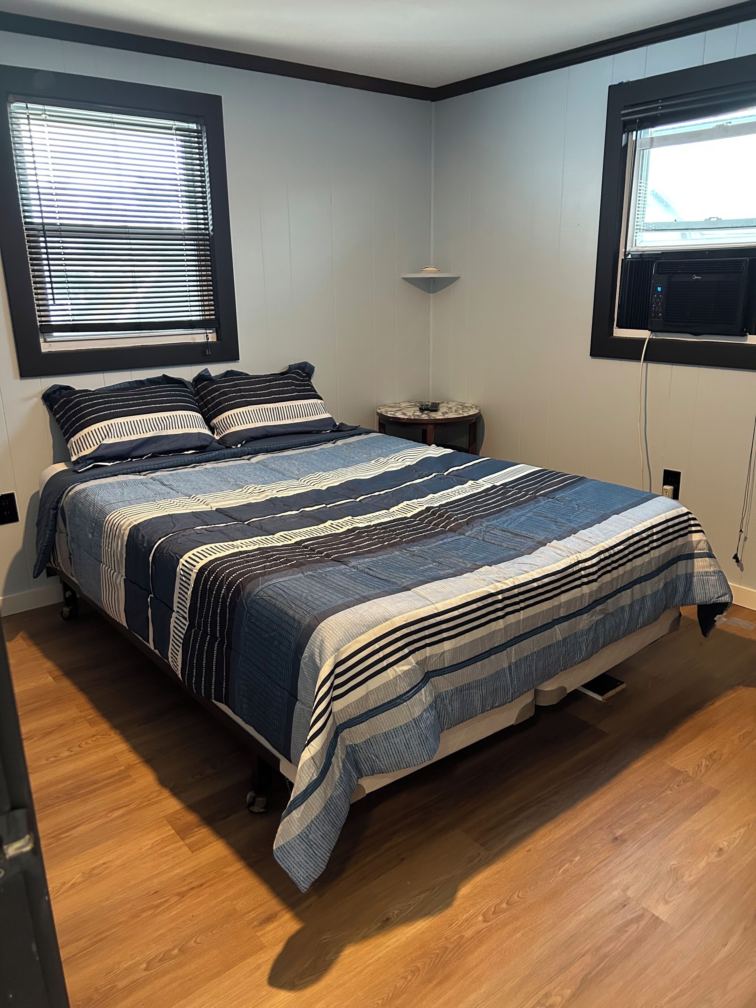 A bedroom with a bed, two windows, a bedside table, and a wall-mounted air conditioner.