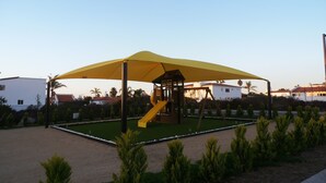 Ein Spielplatz mit einer gelben Rutsche und einem überdachten Spielbereich, umgeben von kleinen Bäumen und Sandboden.