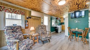Ein gemütliches Zimmer mit Holzbalkendecke, einem an der Wand montierten Fernseher, einem Esstisch mit Stühlen und einem gemusterten Sessel.