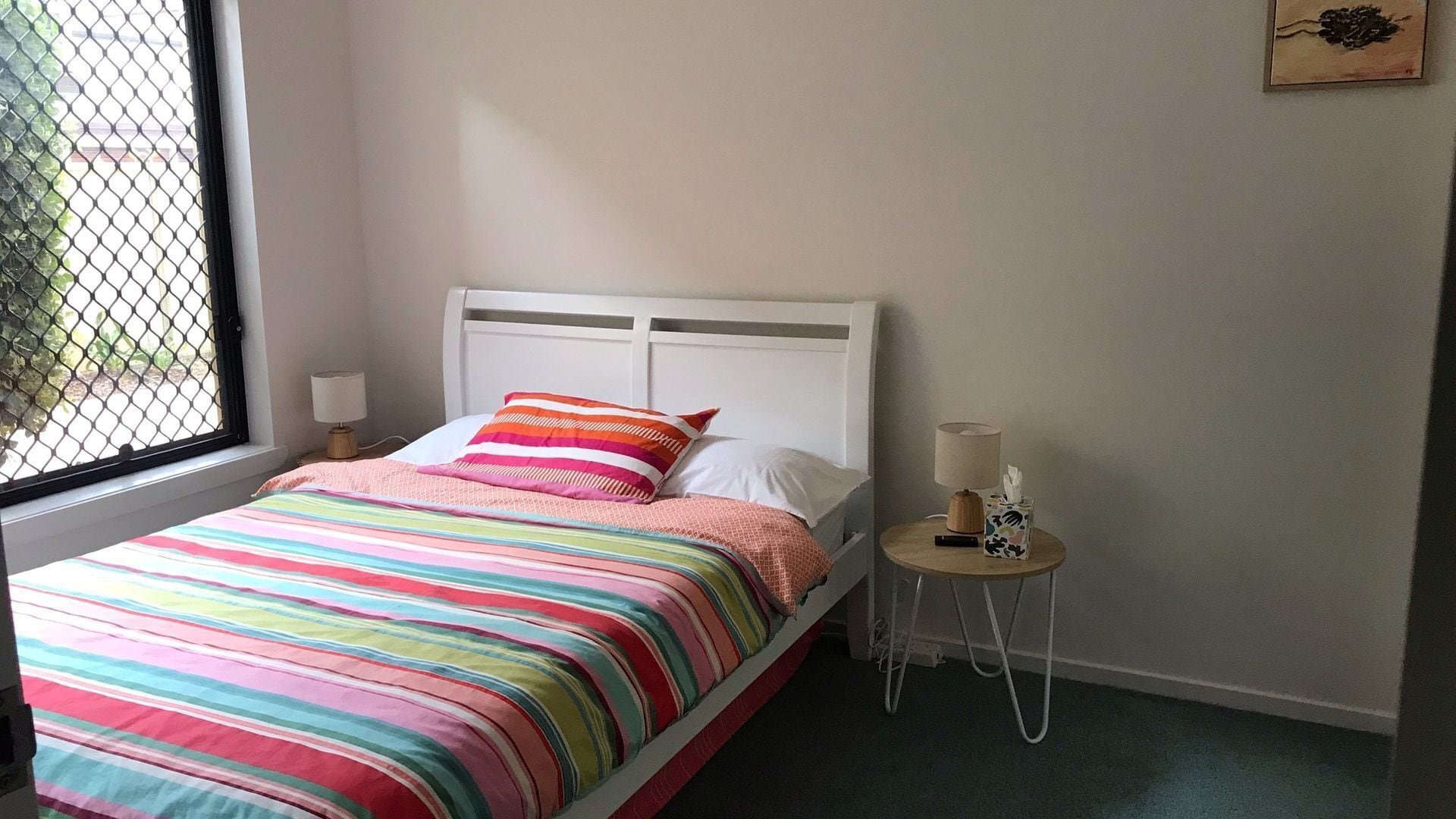 A bed with a colorful striped bedspread and pillows, a bedside table with a lamp and a tissue box, and a window with a metal screen.