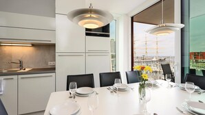 Un espace repas moderne doté d’une grande fenêtre donnant sur un port et sur de grands voiliers. On y trouve des tables à manger blanches, mises avec des assiettes, des verres et des couverts.