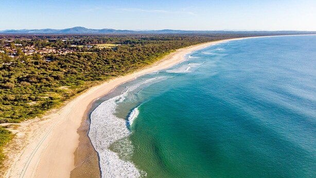 Vue aérienne d’une zone côtière avec une plage de sable, des eaux d’un bleu limpide et un paysage boisé en arrière-plan.