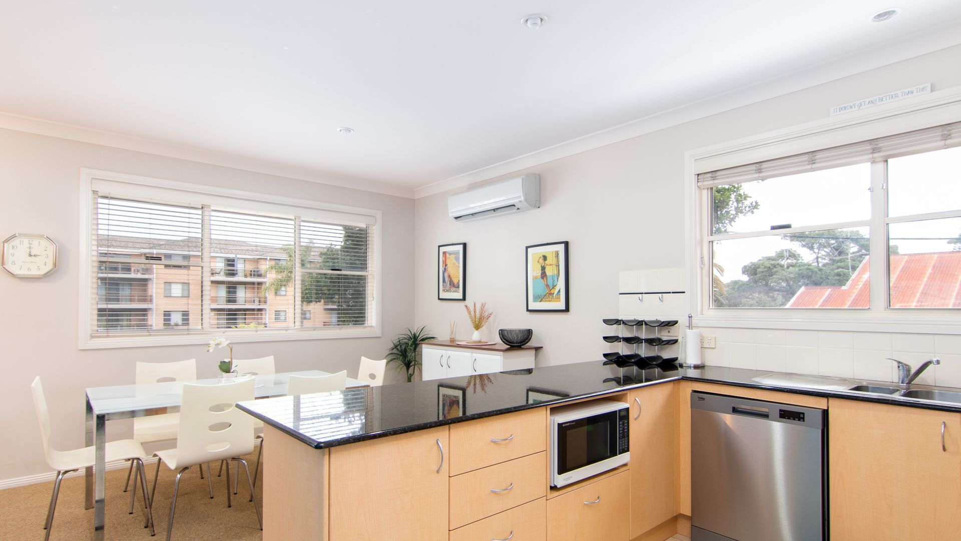 A modern kitchen with a dining area, featuring a stainless steel dishwasher, a microwave, and a sink. There are framed pictures on the wall and a clock on the wall.